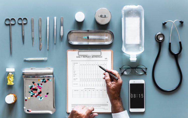 An image of various medical equipment laid out on a blue table such as a stethoscope, glasses, medical scissors, needles, pills and more.